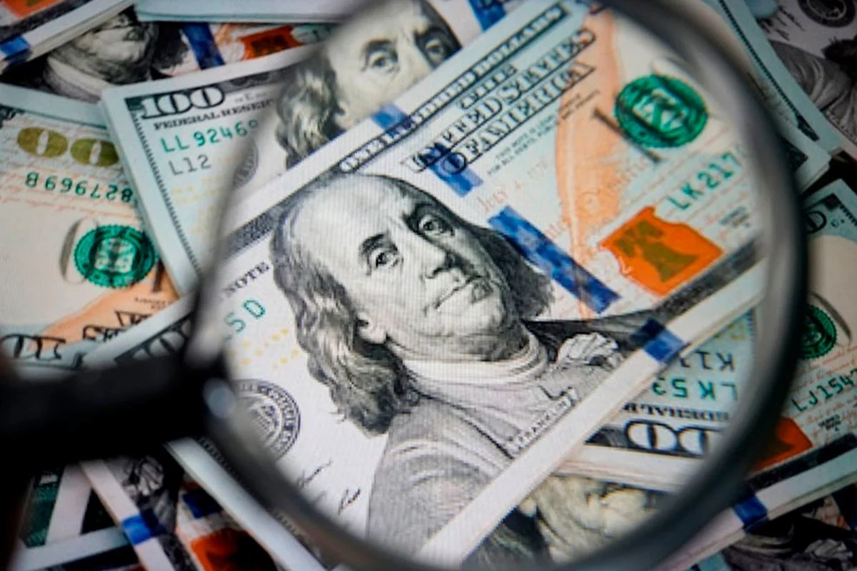 Imagem de notas de dólar, empilhadas umas sobre as outras, com uma lupa sobre elas - Metrópoles