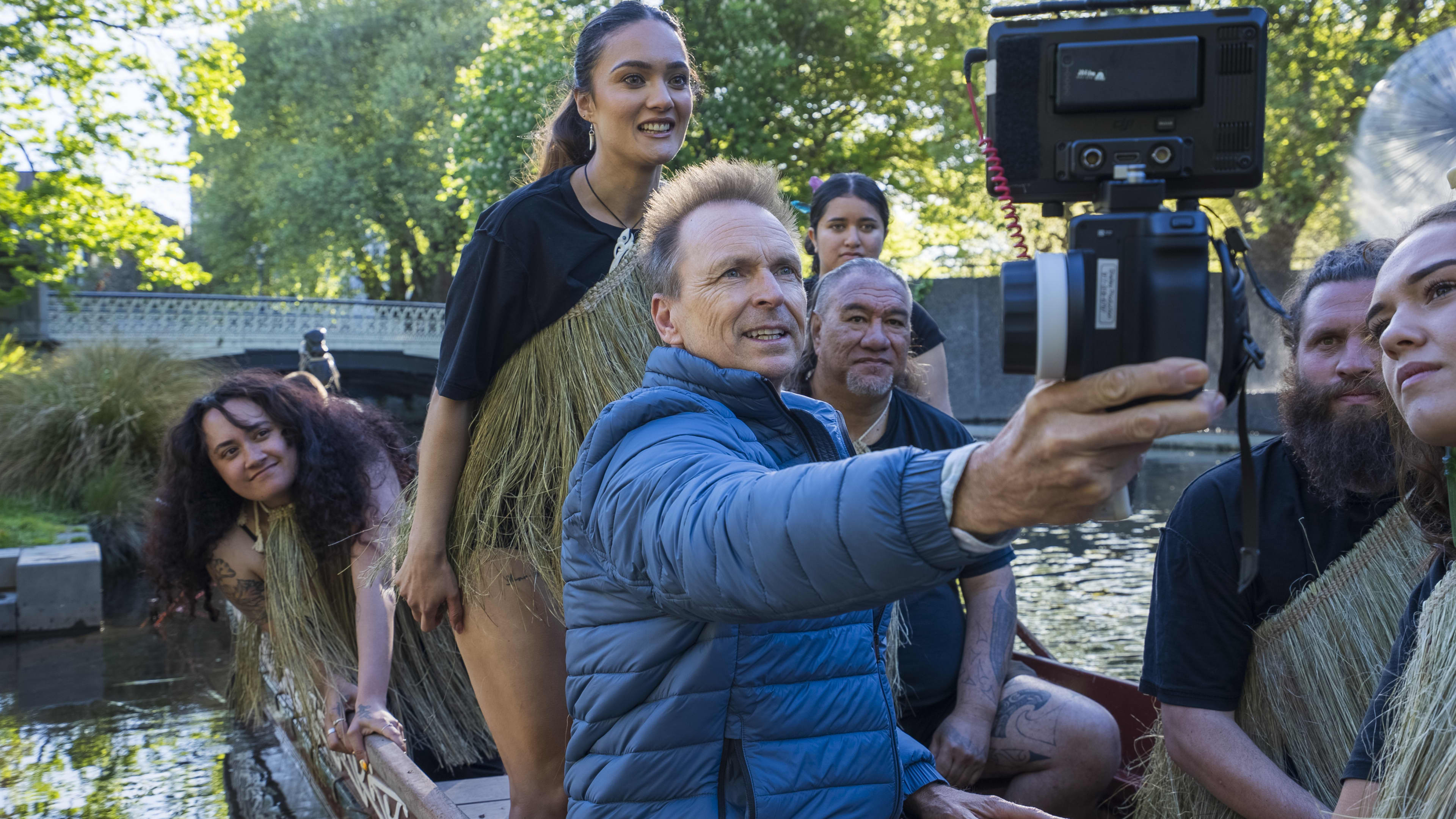 The Amazing Race host Phil Keoghan holds up a camera with fellow Māori Kiwis in a waka at Avon River in Christchurch.